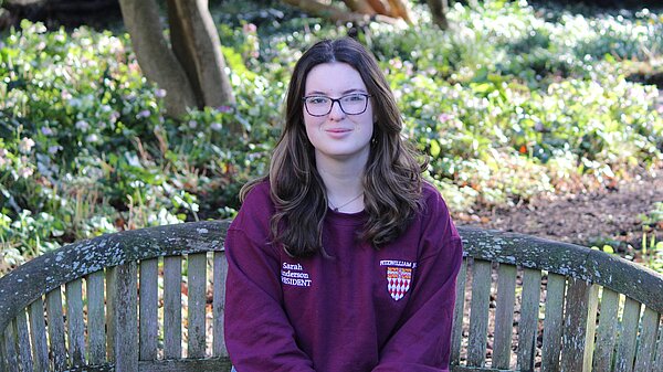 Sarah, a white woman with brown hair wearing a maroon jumper, sits on a bench