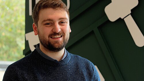 Tom, a white man with brown hair and a beard, wears a blue jumper in Lib Dem HQ